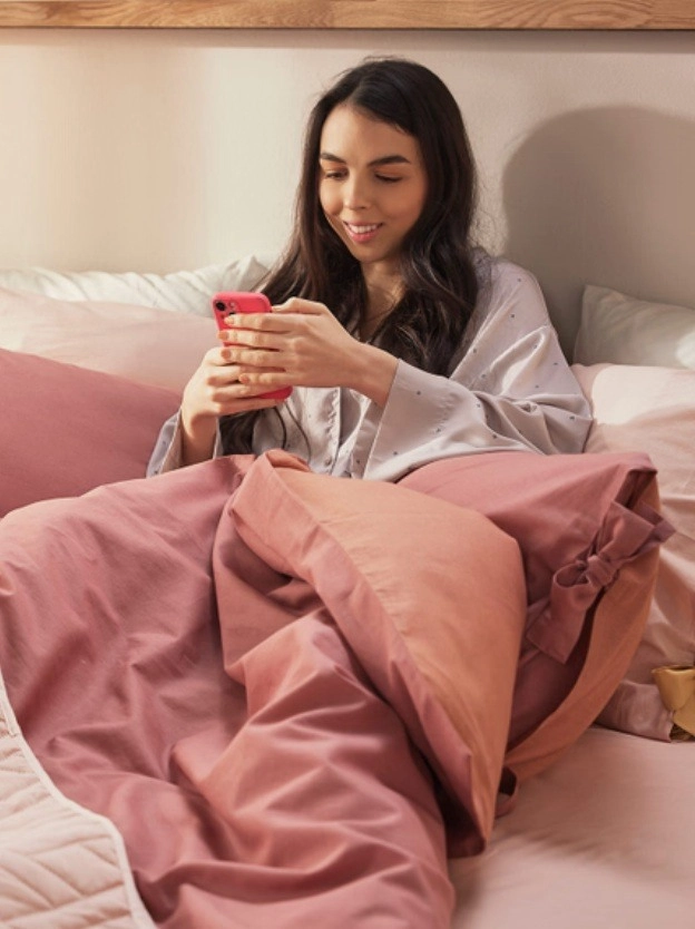 A lány az ágyban fekve mobilozik, szép rózsaszín ágyneműje van