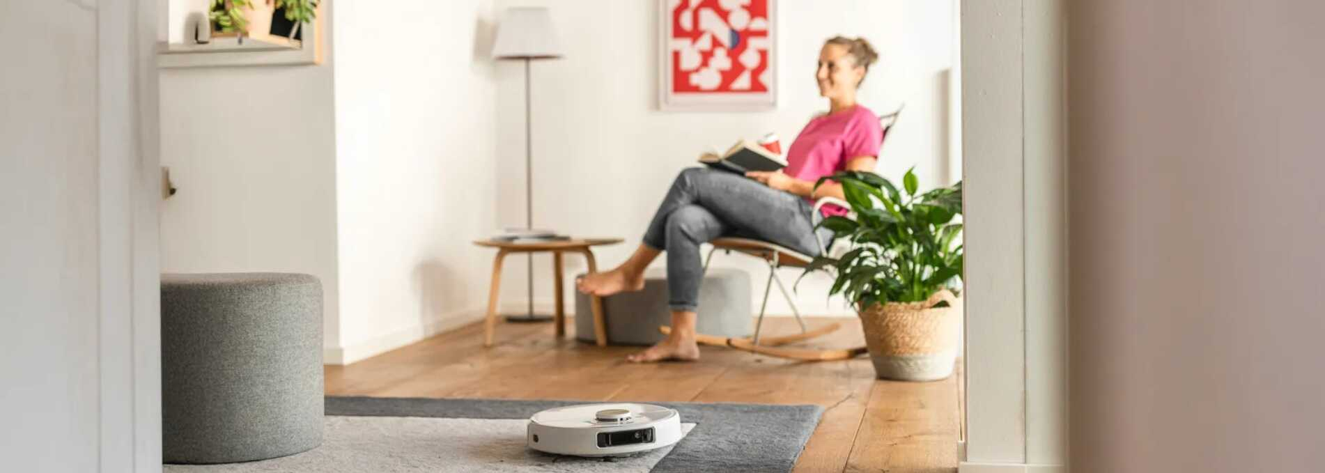 A nő ücsörögve könyvet olvas, míg a robotporszívó takarít