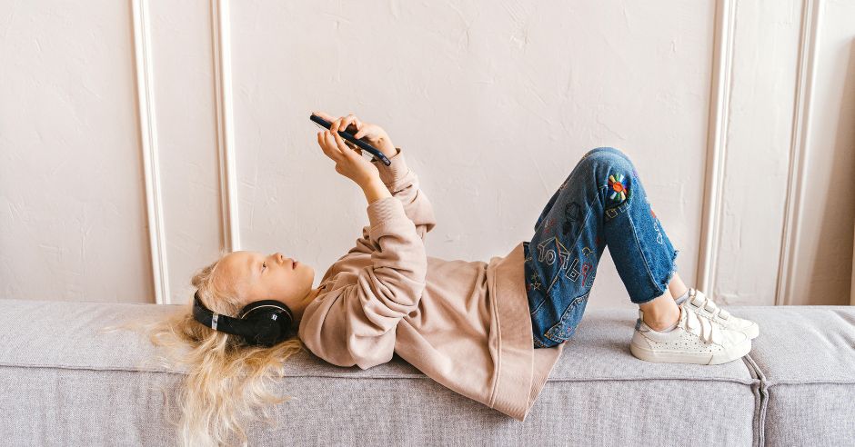 A kislány a kanapén fekve fülhallgatón keresztül hallgat zenét a mobiltelefonjáról.